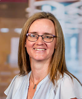 A woman wearing glasses and a blue shirt stands in an office with her arms crossed. AI generated content