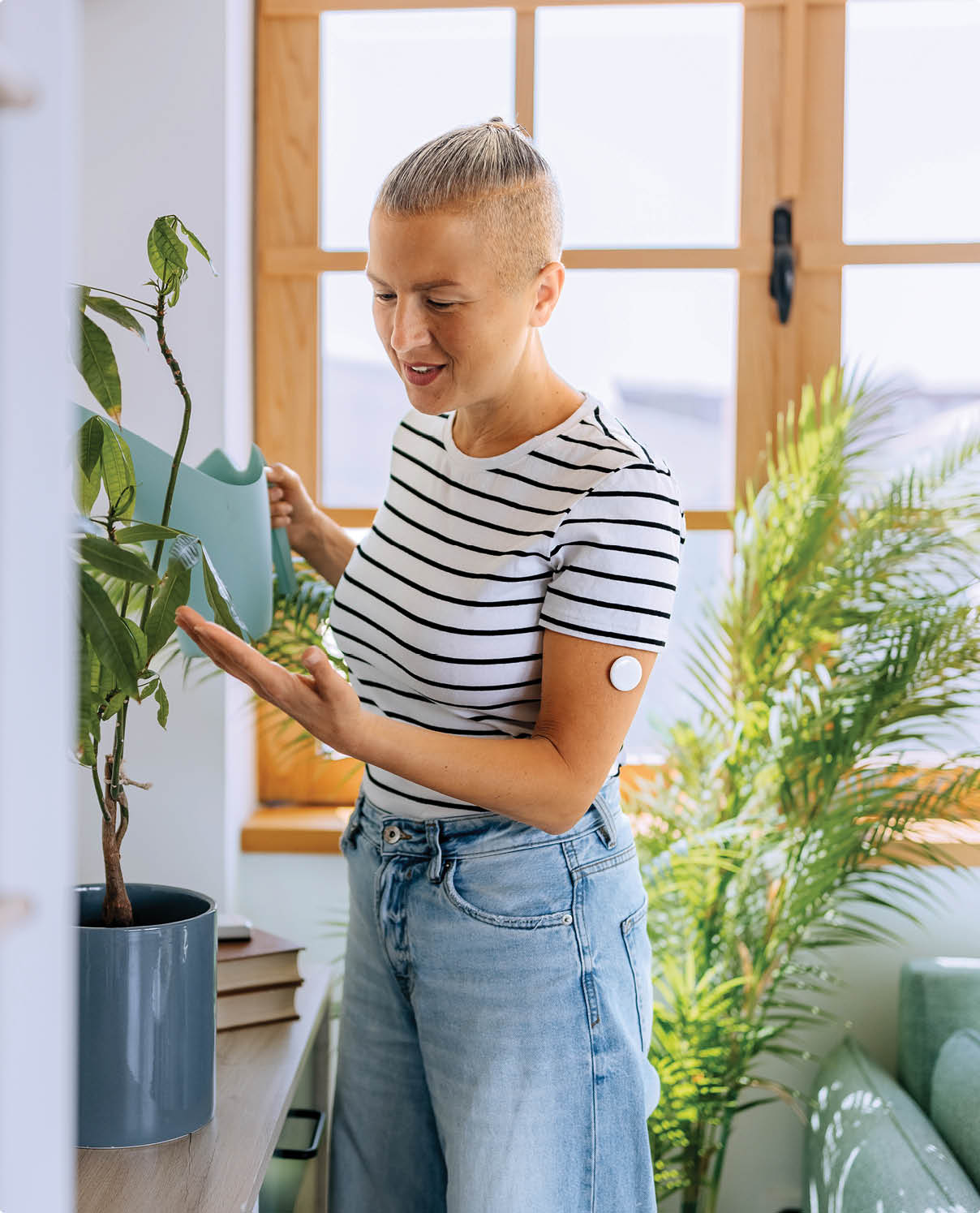 A woman wearing a glucose sensor on her arm tending to her indoor plants in a bright and cozy home. The scene reflects wellness, mindfulness, and everyday health management.